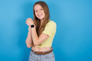 Dreamy young ukranian girl wearing yellow t-shirt over blue backaground with pleasant expression, closes eyes, keeps hands crossed near face, thinks about something pleasant