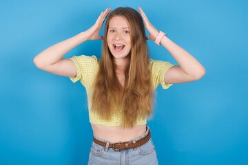 young ukranian girl wearing yellow t-shirt over blue backaground looks with excitement at camera, keeps hands raised over head, notices something unexpected, reacts on sudden news.