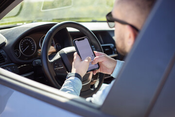 Young bearded man with eyeglasses sitting in his car and reading message on smart phone while waiting in traffic.