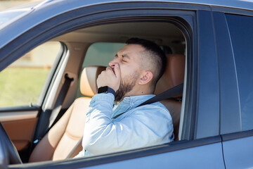 young man looks tired yawning while siting in his car with open window, safety driving concept.