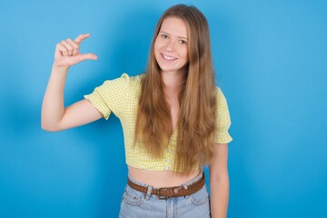 young ukranian girl wearing yellow t-shirt over blue backaground smiling and gesturing with hand small size, measure symbol.