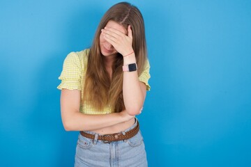 young ukranian girl wearing yellow t-shirt over blue backaground making facepalm gesture while...