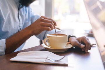 Close-up cropped shot of unrecognizable young adult man mixing hot morning coffee with spoon sitting at desk in cafe on blurred background of window.