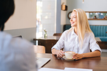 Front view of smiling attractive blonde woman sitting in front of unrecognizable young man at table of cafe and holding mug of hot coffee, looking away. Romantic date in cafe, couple enjoying time.