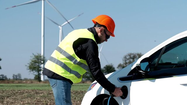 Windmills charge white electric car in field. Professional engineer removes plug from ev socket against rotating propellers at rural site