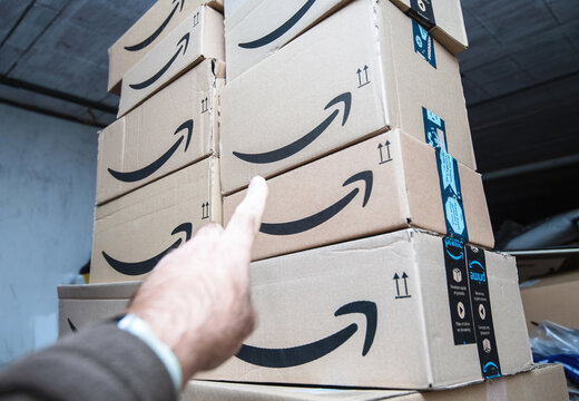 Paris, France - Oct 20, 2021: POV male hand showing to large pile stack in warehouse distribution center garage of Amazon Prime parcel goods boxes with smiling amazon arrow