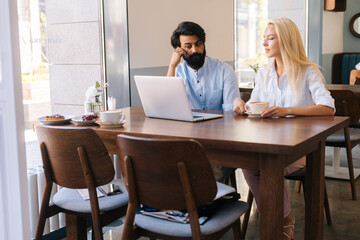 Portrait of young focused businesswoman and businessman working together using laptop sitting at cafe by window and drinking coffee. Two business partners or colleagues working in coffee shop.