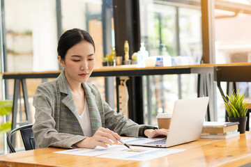 Asian businesswoman holding pen pointing to the graph to analyze marketing plan with calculator and laptop on wooden desk in office Finance and Accounting Concepts