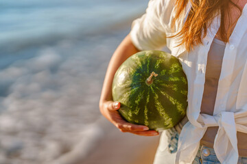 A young woman in a white shirt carries a watermelon in her hands, walking along the sea on the beach. Summer picnic on the beach