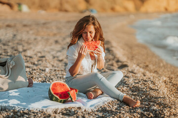 A young woman in a white shirt and jeans is sitting on the beach and eating a watermelon. The concept of a quiet summer evening and a picnic with fruit