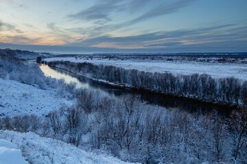 A beautiful winter view from the hill to the river with frosty trees growing along the banks. Winter landscape at sunset