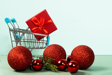 Beautiful red gift box wrapped in ribbons with red Christmas balls in a shopping cart. Concept of Christmas and New Year online shopping around the world. Front view on a light background.