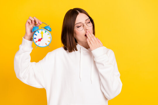 Photo of exhausted sleepy lady hold alarm clock yawn wear glasses white hoodie isolated yellow color background