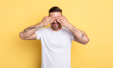 young handsome man smiling and feeling happy, covering eyes with both hands and waiting for...