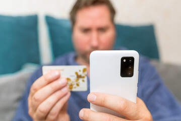 Young man buying fashion items through the Internet on cellphone by using credit card.Online Payment. Frontal portrait of man holding debit card and using smartphone for shopping at home