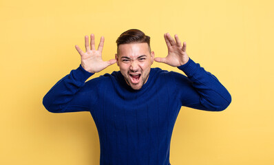 young handsome man screaming in panic or anger, shocked, terrified or furious, with hands next to...