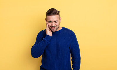 young handsome man holding cheek and suffering painful toothache, feeling ill, miserable and...