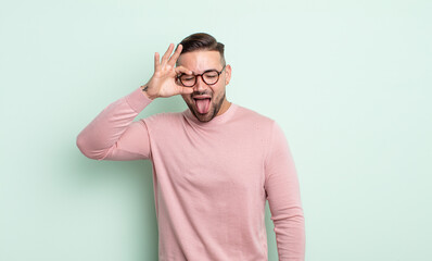 young handsome man smiling happily with funny face, joking and looking through peephole, spying on...