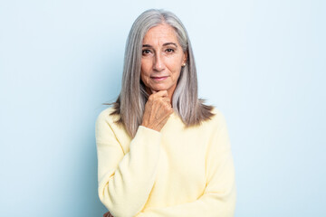 middle age gray hair woman looking happy and smiling with hand on chin, wondering or asking a...