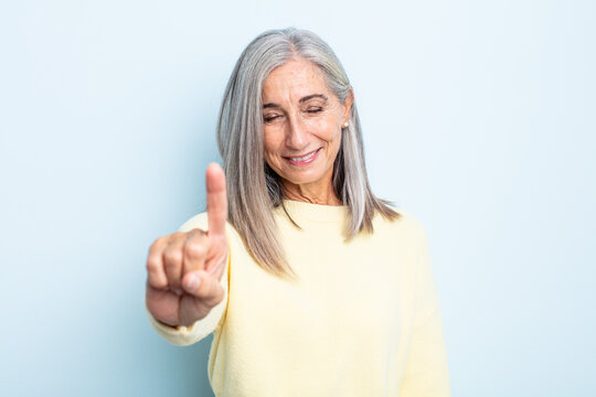 middle age gray hair woman smiling and looking friendly, showing number one or first with hand forward, counting down