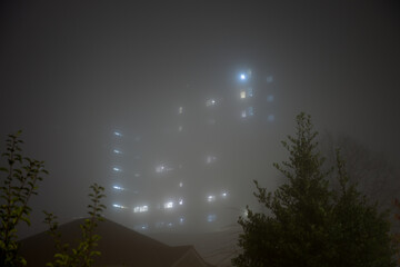 Eerie sky scraper hospital lights shine through very thick fog.