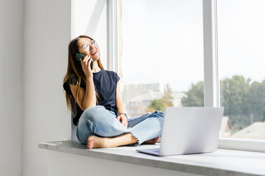 Portrait of charming woman talking on the smartphone sitting on the window cill at home.