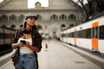 Young woman waiting for the train standing at the railway station. Beautiful girl using the phone while waiting for the train.