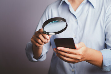 Search online concept. Close-up of the hand of a businesswoman wearing a blue shirt holding a magnifying glass looking at her phone while standing in a gray background in the studio