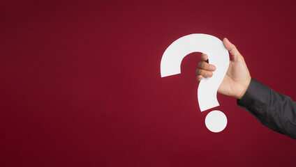 Hand of a woman in a black shirt holding a white question mark paper while standing with a red background in the studio. Close-up photo. Space for text