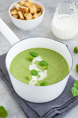 Broccoli cream soup with fresh basil and in a white saucepan on a gray concrete background.