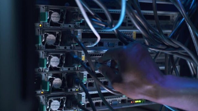 Engineer in data center. IT specialist black man hand switches cable to connect operating spbas equipment on rack in semi-dark server room closeup