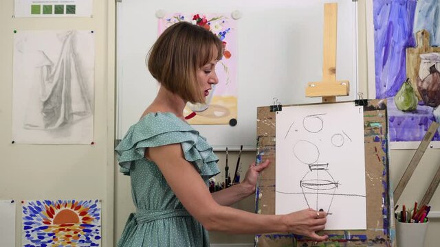 Painter woman sketch picture on easel, teacher look at camera and talk about art Spbas. Portrait creative blogger stand in workshop and give lesson, explain theory. Concept education, tutor, online