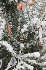 Modern christmas ornaments hanging on snowy pine tree branch. Decorated christmas tree with stylish baubles in snow outdoors. Winter holidays in countryside. Merry Christmas! Space for text