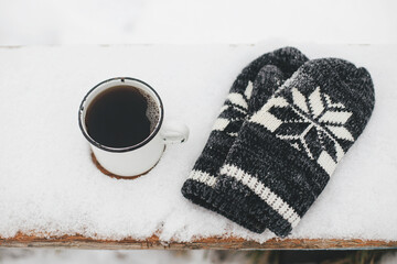 Warm cup of tea and cozy knitted mittens in snow on wooden bench in winter park. Hiking and traveling in cold season. Wanderlust. Space for text. Winter time