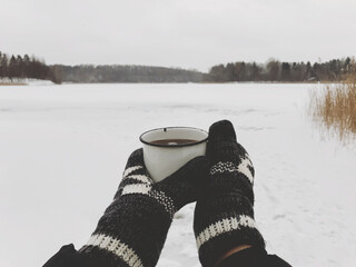 Hands in cozy gloves holding warm cup of tea on background of  snow lake in winter. Hiking and traveling in cold winter season. Warm drink. Space for text. Wanderlust