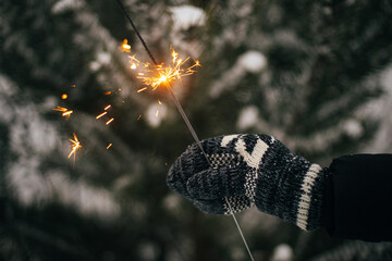 Hand in cozy glove holding burning sparkler on background of pine tree branches in snow. Happy New Year! Atmospheric magic moment. Woman hand with glowing firework in evening. Happy Holidays!