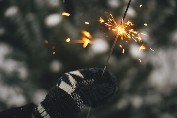 Happy New Year! Hand in cozy glove holding burning sparkler on background of pine tree branches in snow. Atmospheric magic moment. Woman hand with glowing firework in evening. Happy Holidays!