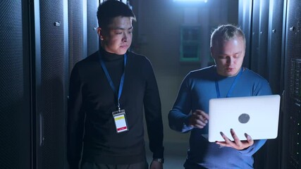 Male data engineers talking and working with computer while walking in server room spbas. Close-up view of two young men discuss business with smiles and check complex equipment, hold laptop in hand