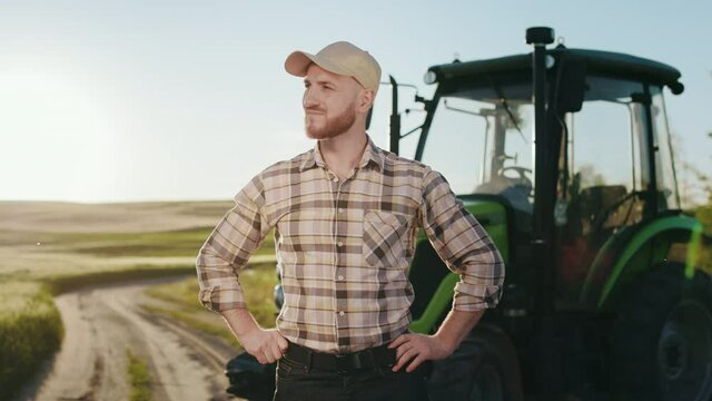 A young bearded farmer is putting on a cap and keeping his hands on his belt. He is standing in the middle of a field near a tractor. Sunset in the background. 4K