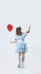 Back view of young girl wearing Halloween dress, costume of movie character standing with red balloon in hand isolated over blue background