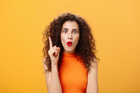 I got excellent plan. Enthusiastic emotive and excited female with curly hairstyle in orange cropped top raising index finger in eureka gesture folding lips popping eyes adding amazing suggestion