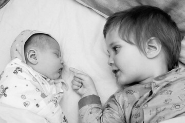 Caucasian toddler boy lying on bed opposite his newborn swaddled sister looking at her and...