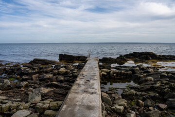 Fototapeta na wymiar Rocky shoreline with a concrete jetty. Blue water and a blu sky in the background. Picture from Skalderviken, southern Sweden