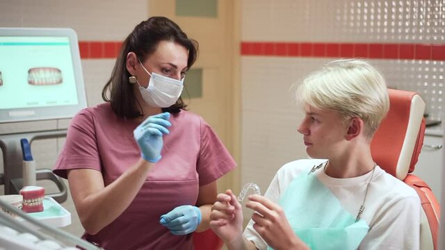 Orthodontist and teenage client at appointment. spbd Brunette doctor in pink uniform gives aligners to blond schoolboy patient in contemporary hospital office