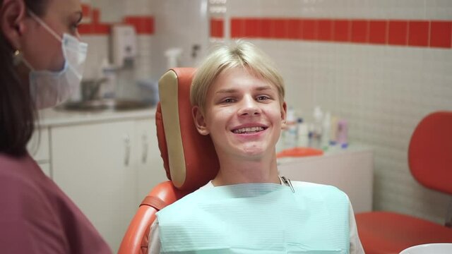 Orthodontist consults teenager patient. Blond teen boy smiles showing transparent aligners spbd on teeth at appointment with doctor in clinic office
