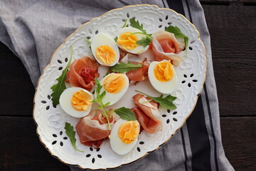 Italian salad. Boiled eggs with smoked ham and arugula on white plate. Dark wooden background