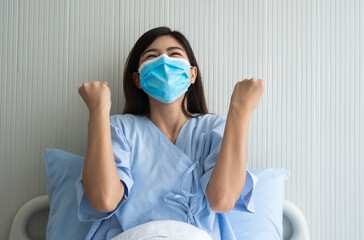 Happy Asian female patient wearing a mask, lies on the bed, and I raised an arms and was delighted for show confidence in treatment. Concept of believe in treatment And insurance coverage