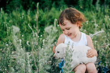 Little cute caucasian girl smiling sitting in high grass and holding white puppy in her lap, looking at pet with love. Blurred green background. Horizontal shot, front view. Pet love concept.