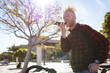 Happy albino african american man with dreadlocks in park with bike talking on smartphone