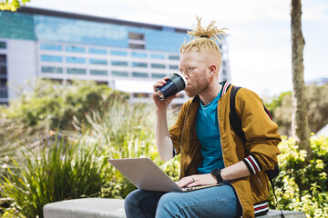Thoughtful albino african american man with dreadlocks sitting in park drinking coffee, using laptop